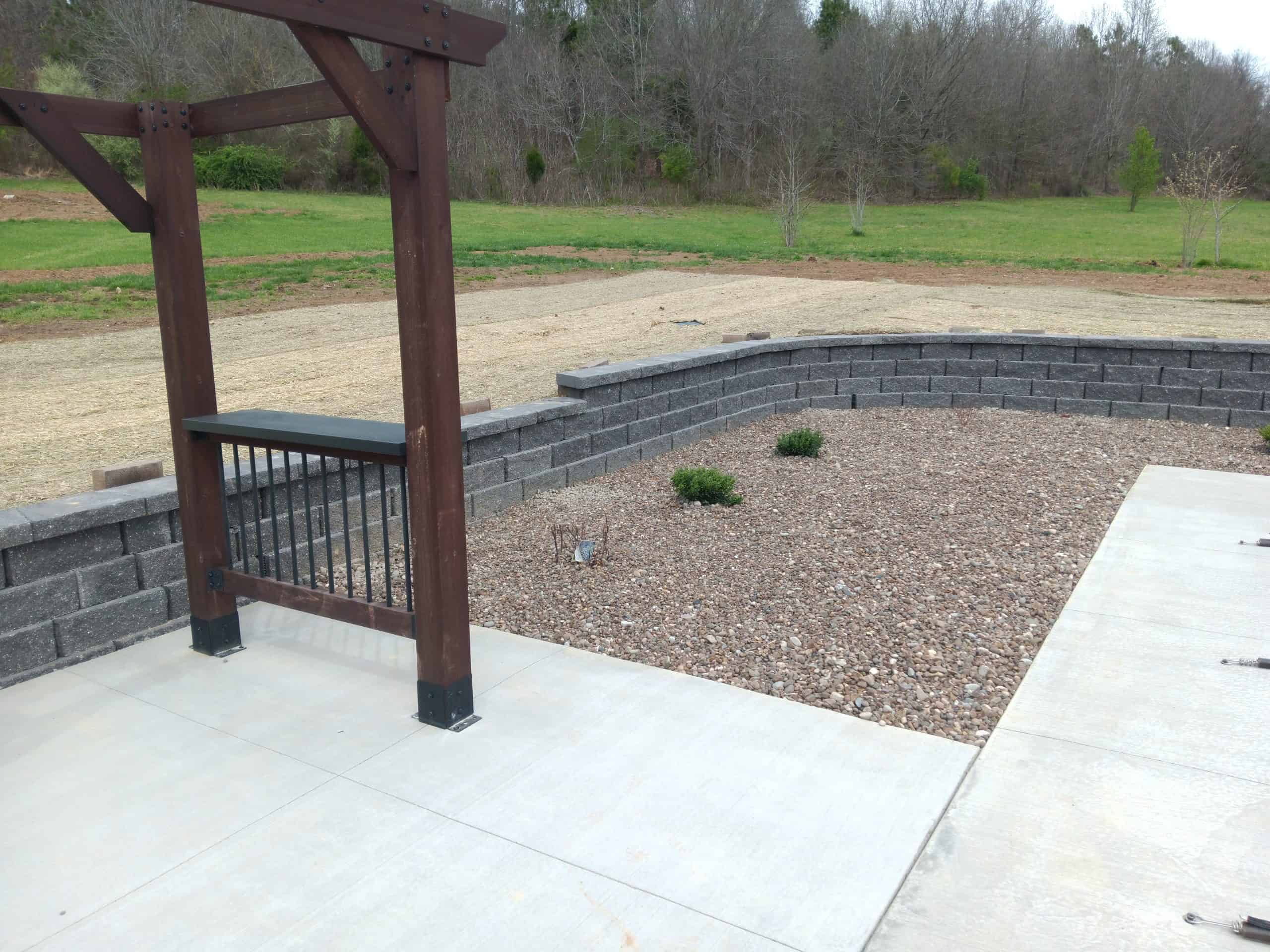 a hardscaped patio bordered by retaining walls with a wooden pergola overlooking a tree line
