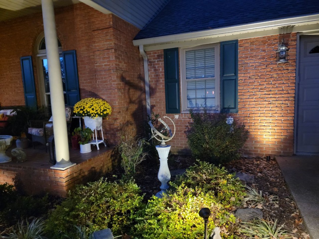 GoPerfection-CentralCity-LandscapeLighting Front porch of a brick house with a sundial statue, bright yellow flowers, and outdoor lighting illuminating the landscape.