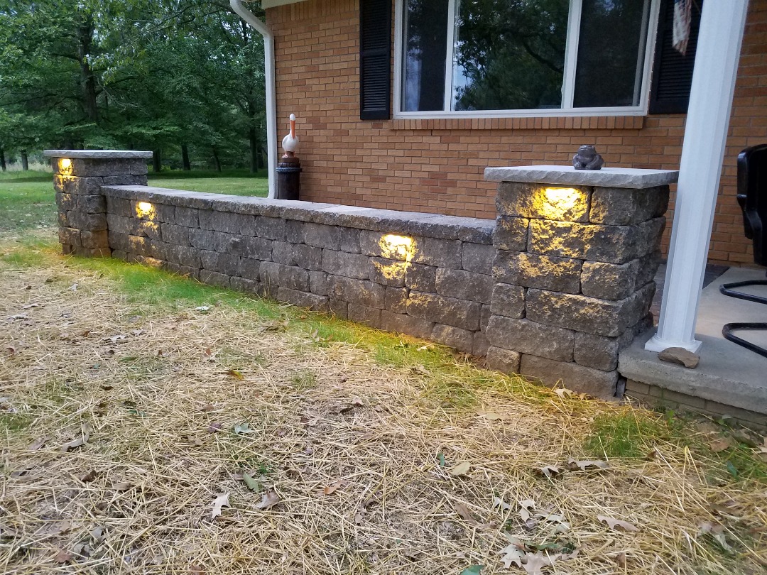 Outdoor wall lights highlight a stone retaining wall in front of a brick house, enhancing the patio entrance with a warm ambiance.