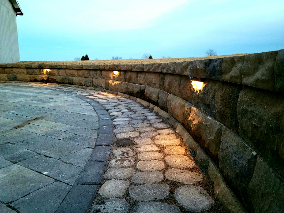 Outdoor wall lights highlight a stone retaining wall in front of a brick house, enhancing the patio entrance with a warm ambiance.