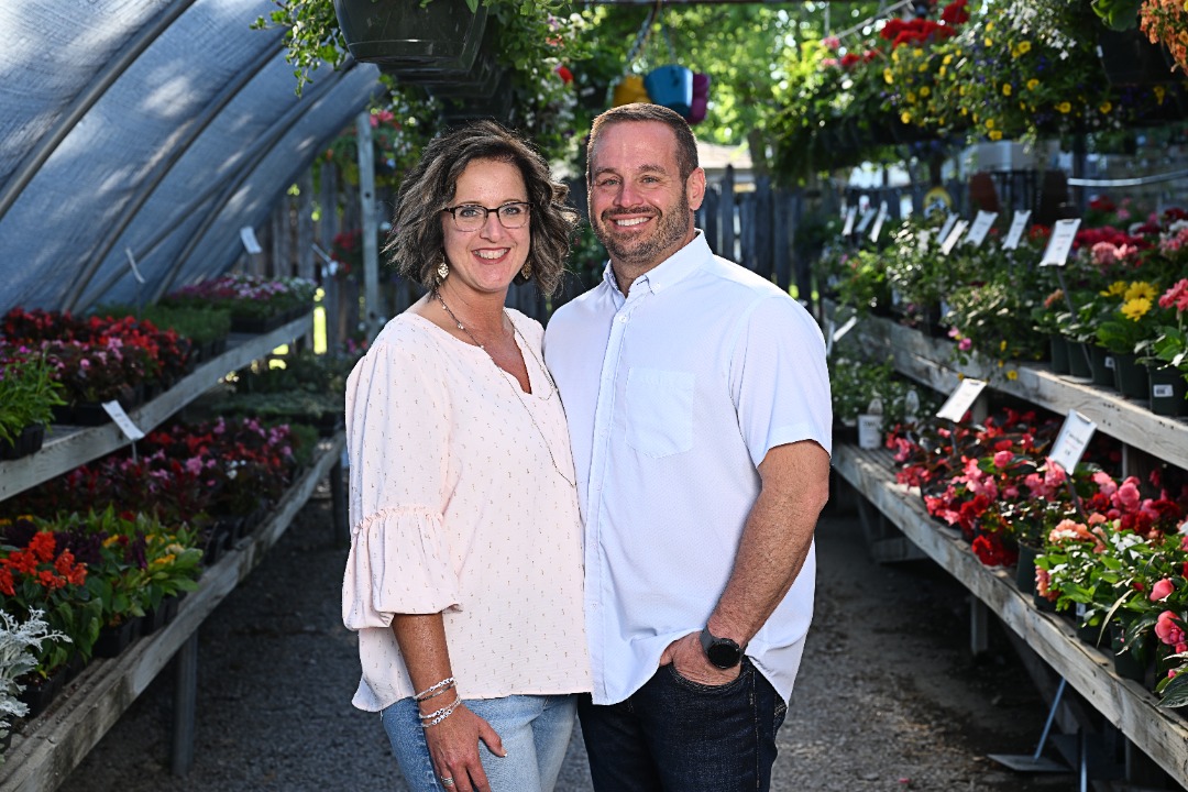 Perfection-About Us - 4 Chris Vinent and his wife stand smiling inside a greenhouse, surrounded by vibrant flowers and plants at their family-owned business.