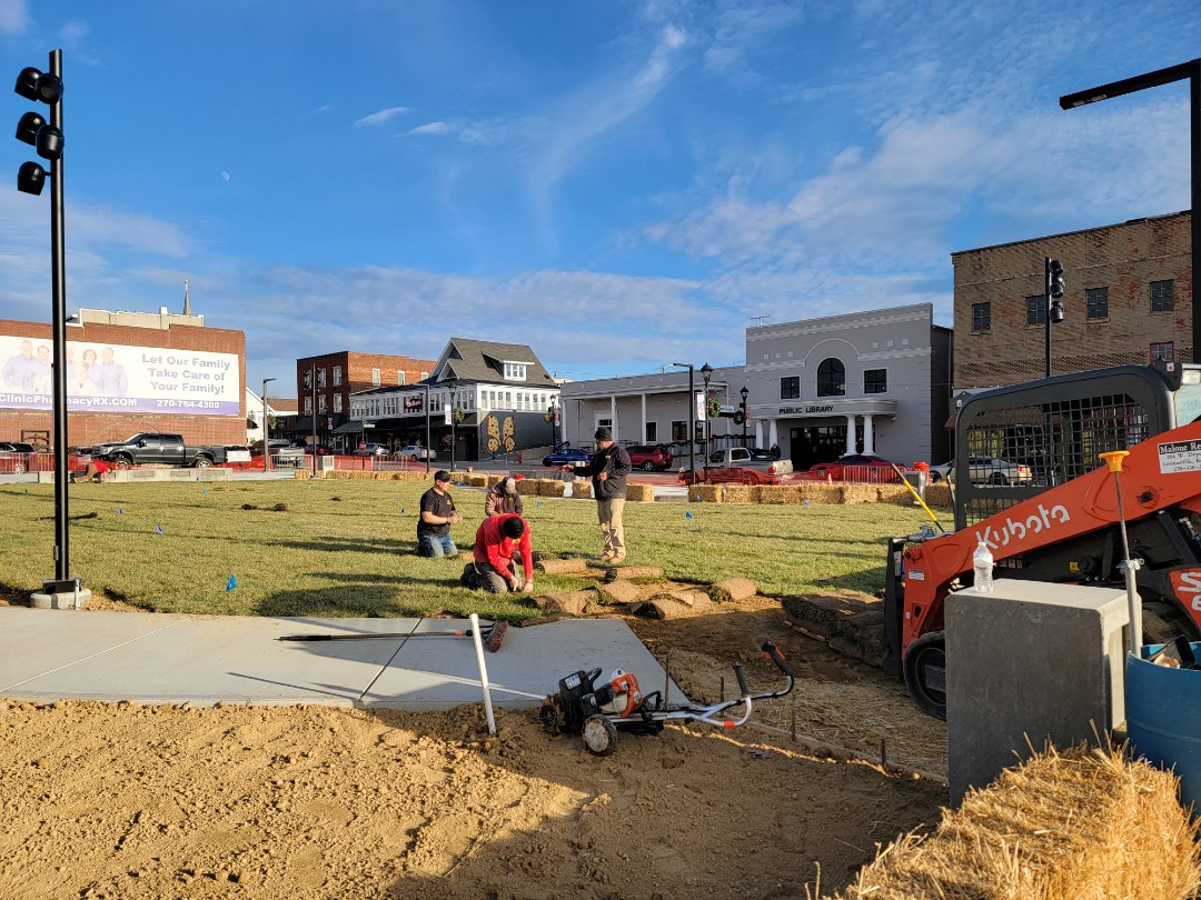 Perfection-About Us - 5 A landscaping crew installs sod on a public lawn, with a skid steer and concrete pathway under construction in a downtown area.