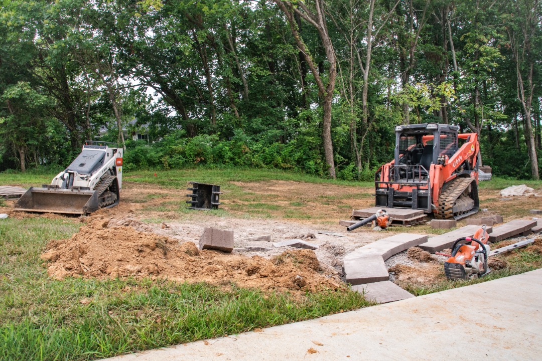 Skid steers and construction tools on a dirt lot with curved pavers being installed for a patio near a wooded area.