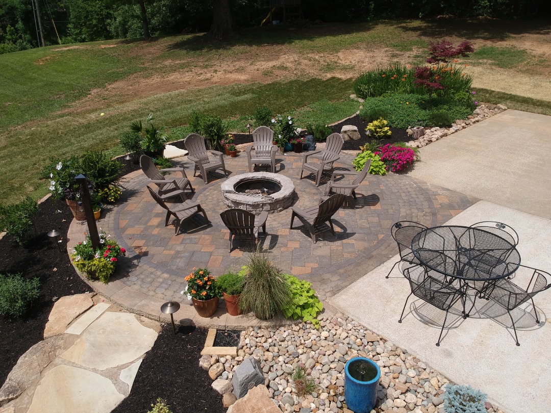 Finished circular paver patio with fire pit, Adirondack chairs, surrounding flowers, and metal dining set on concrete.