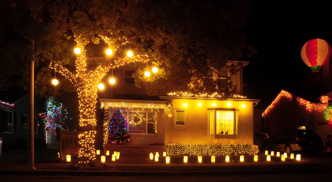 Cozy house with glowing Christmas lights on tree and roofline, lit lanterns lining the path, and a decorated tree inside the window.<br />
