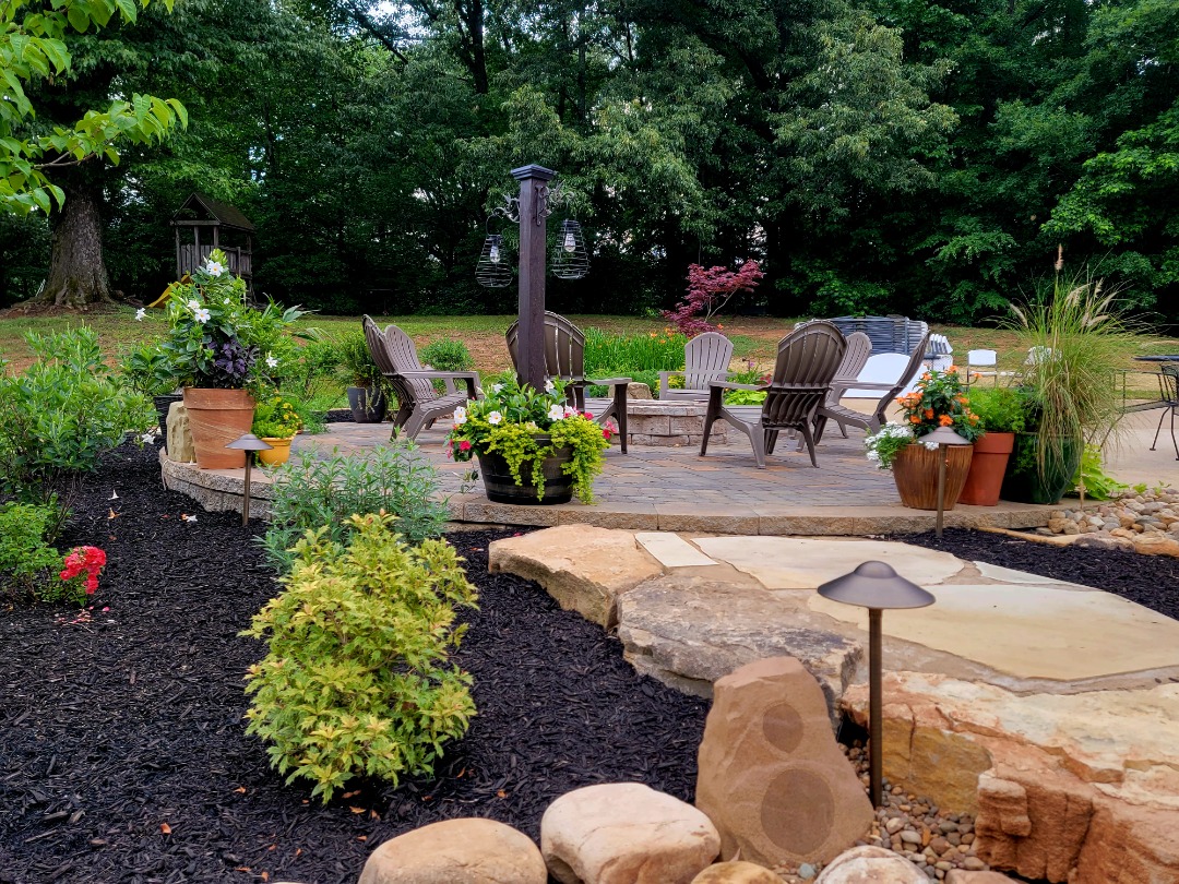 Close-up of a landscaped yard with river rocks, potted plants, stone path, and vibrant greenery beside a red brick house