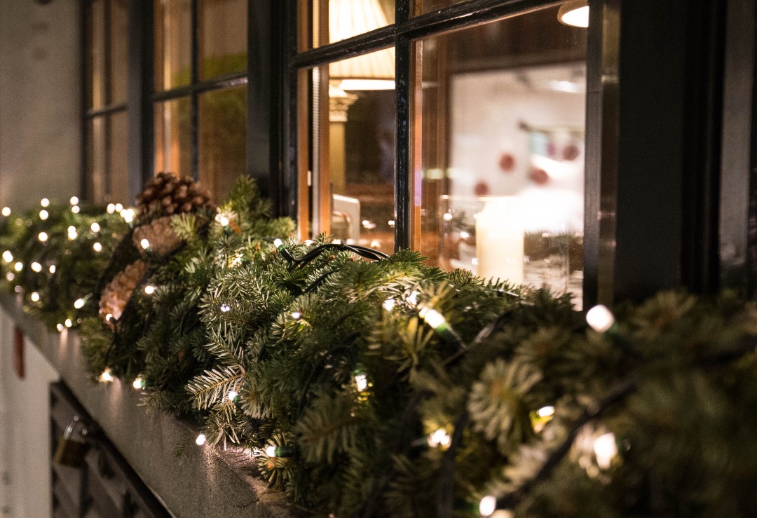 Close-up of holiday garland with pinecones and string lights on a windowsill, reflecting warm indoor lighting through glass panes.
