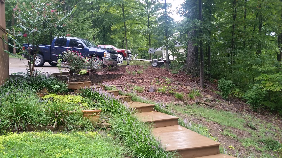 Wooden garden steps lead uphill through lush landscape with flowering bushes, parked trucks, and tall forest backdrop.
