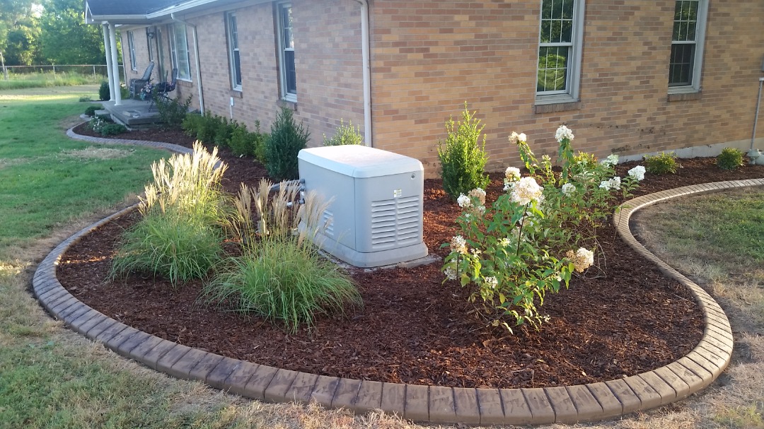 Mulched garden bed with flowering shrubs and backup generator along brick house with curved concrete landscape edging.
