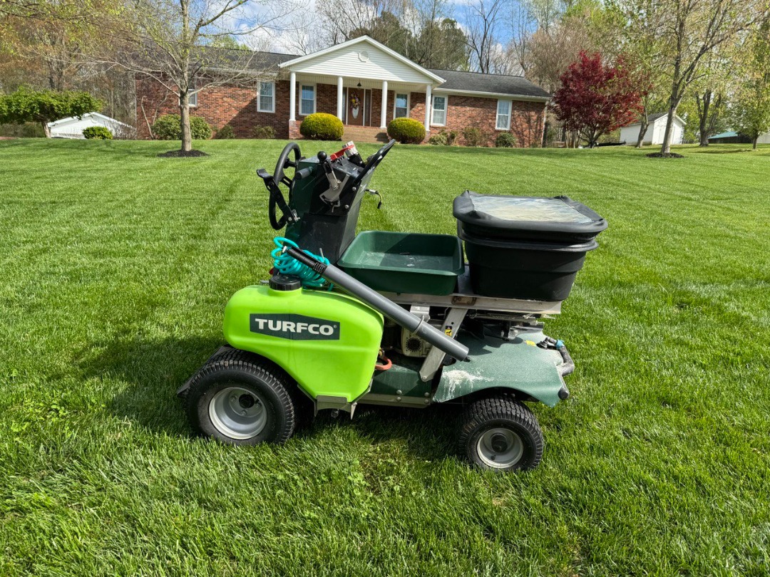 Turfco lawn treatment machine parked on vibrant green lawn in front of brick home with symmetrical landscape design.