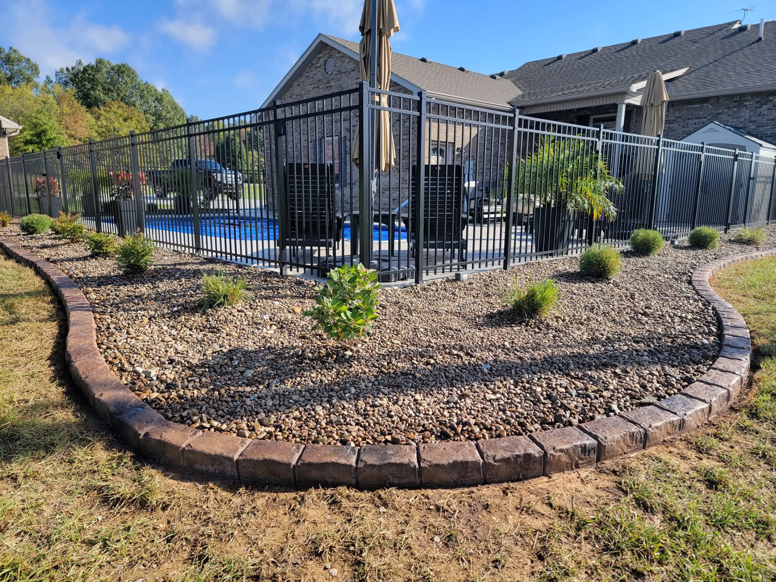 Rock landscaping with brick border and young shrubs lining a black metal fence around a backyard pool in a suburban setting.