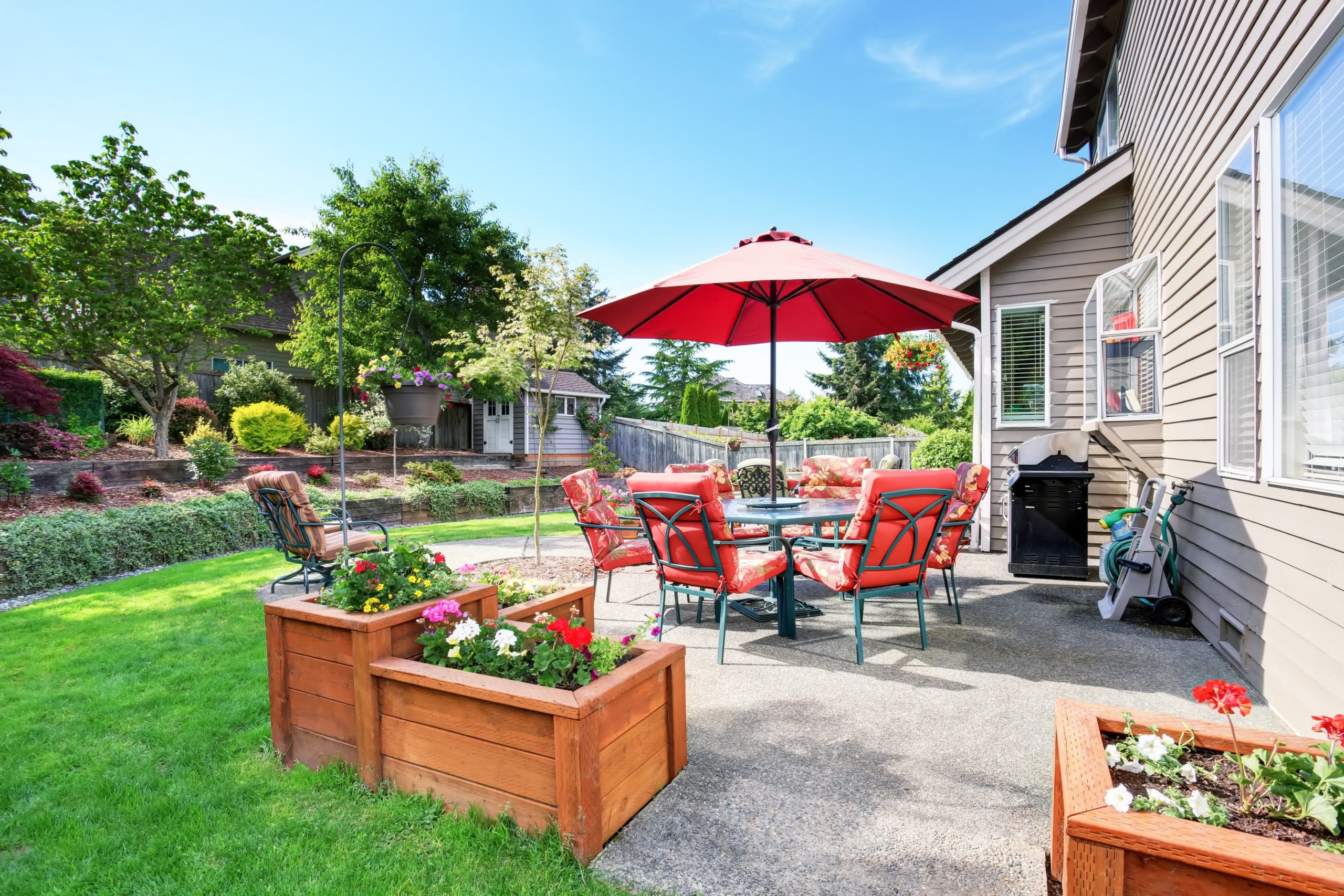 Concrete patio with red umbrella, cushioned chairs, wooden planters, and lush landscaping in a colorful backyard garden.
